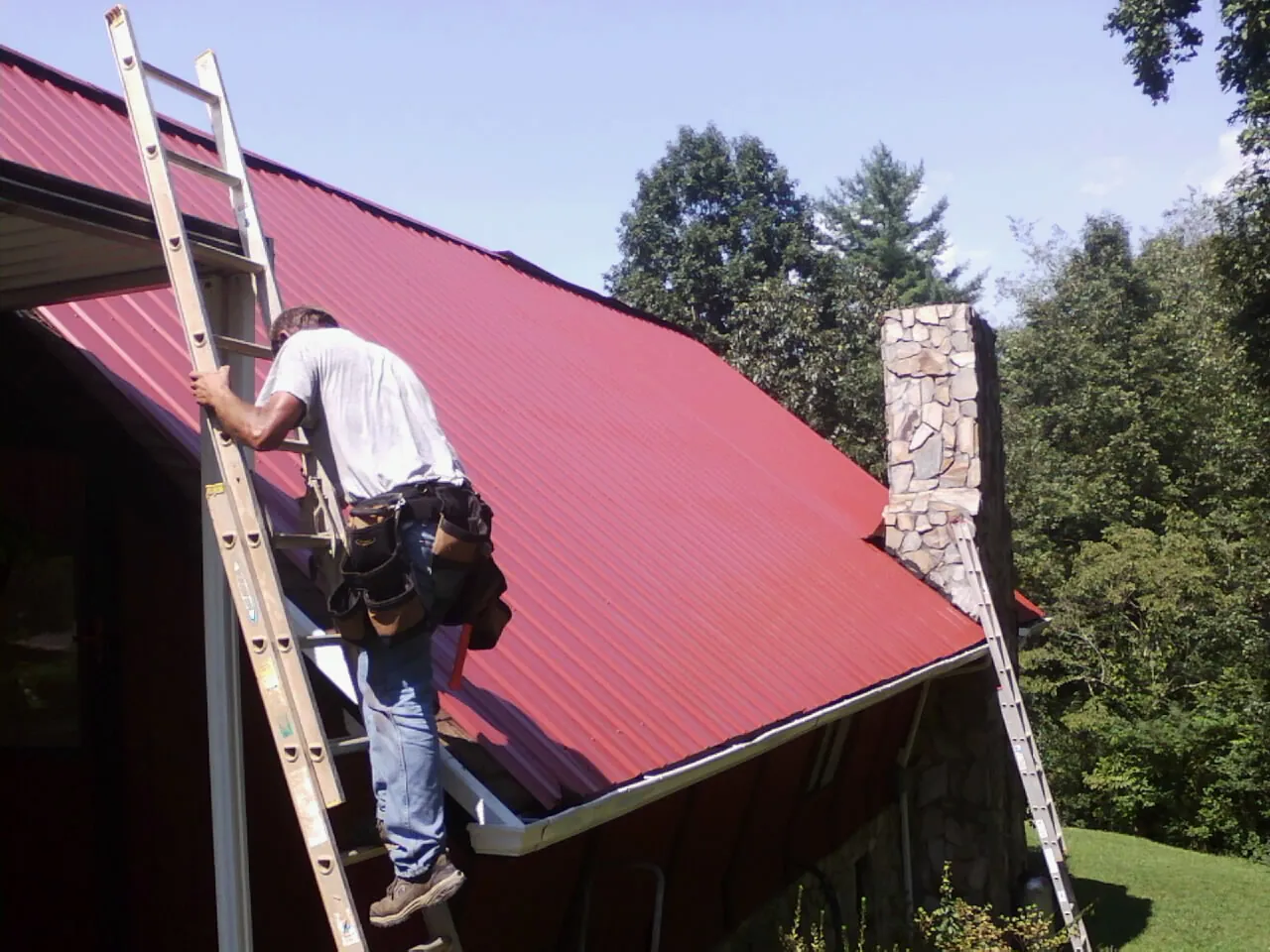 Roofer on a ladder beside a red metal roof and stone chimney — Soffit & Fascia Repair by Roofing Brothers in Mohler